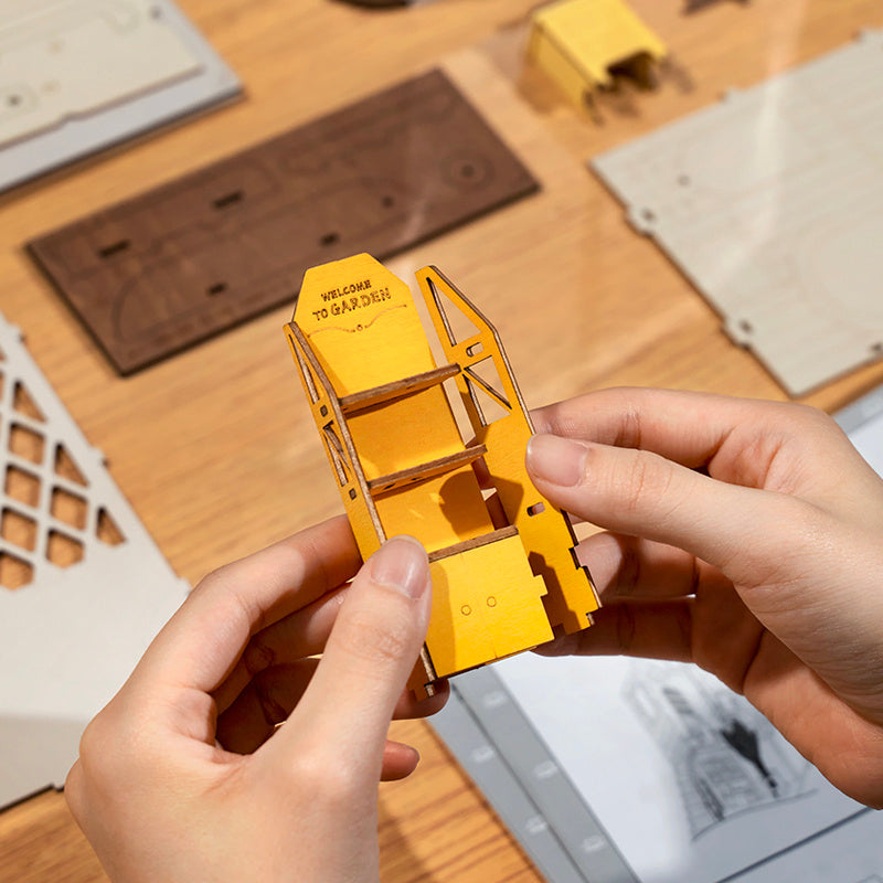 Hands assembling the Rolife Garden House Book Nook Kit with detailed craft pieces on a wooden table